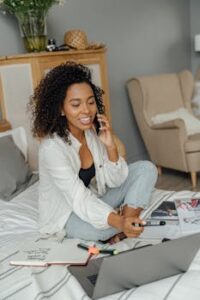 Smiling woman with curly hair talking on phone while working on laptop in cozy bedroom setting.